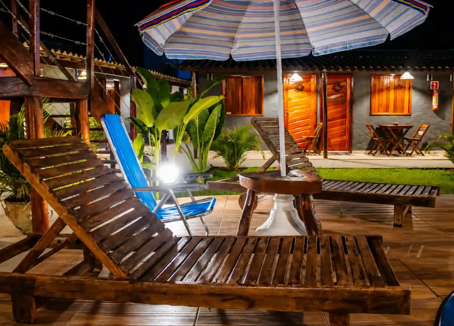 Wooden sun loungers and a parasol on the lit pool deck at night, with guest room doors behind